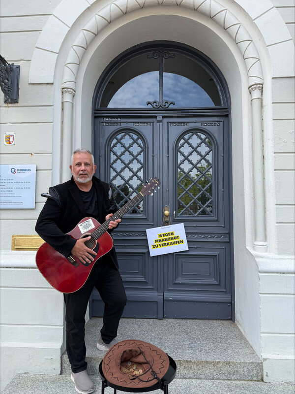 Das Bild zeigt B�rgermeister Saba vor dem Oldenburger Rathaus. Er versucht als Stra�ens�nger Geld f�r die Stadt zu sammeln. Am Rathaus h�ngt ein Schild �zu verkaufen�.
Das Bild will mit einem Augenzwinkern auf die schwierige Finanzlage der Kommunen hinweisen.
