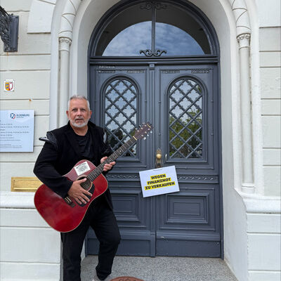 Das Bild zeigt B&uuml;rgermeister Saba vor dem Oldenburger Rathaus. Er versucht als Stra&szlig;ens&auml;nger Geld f&uuml;r die Stadt zu sammeln. Am Rathaus h&auml;ngt ein Schild &raquo;zu verkaufen&laquo;.
Das Bild will mit einem Augenzwinkern auf die schwierige Finanzlage der Kommunen hinweisen.
